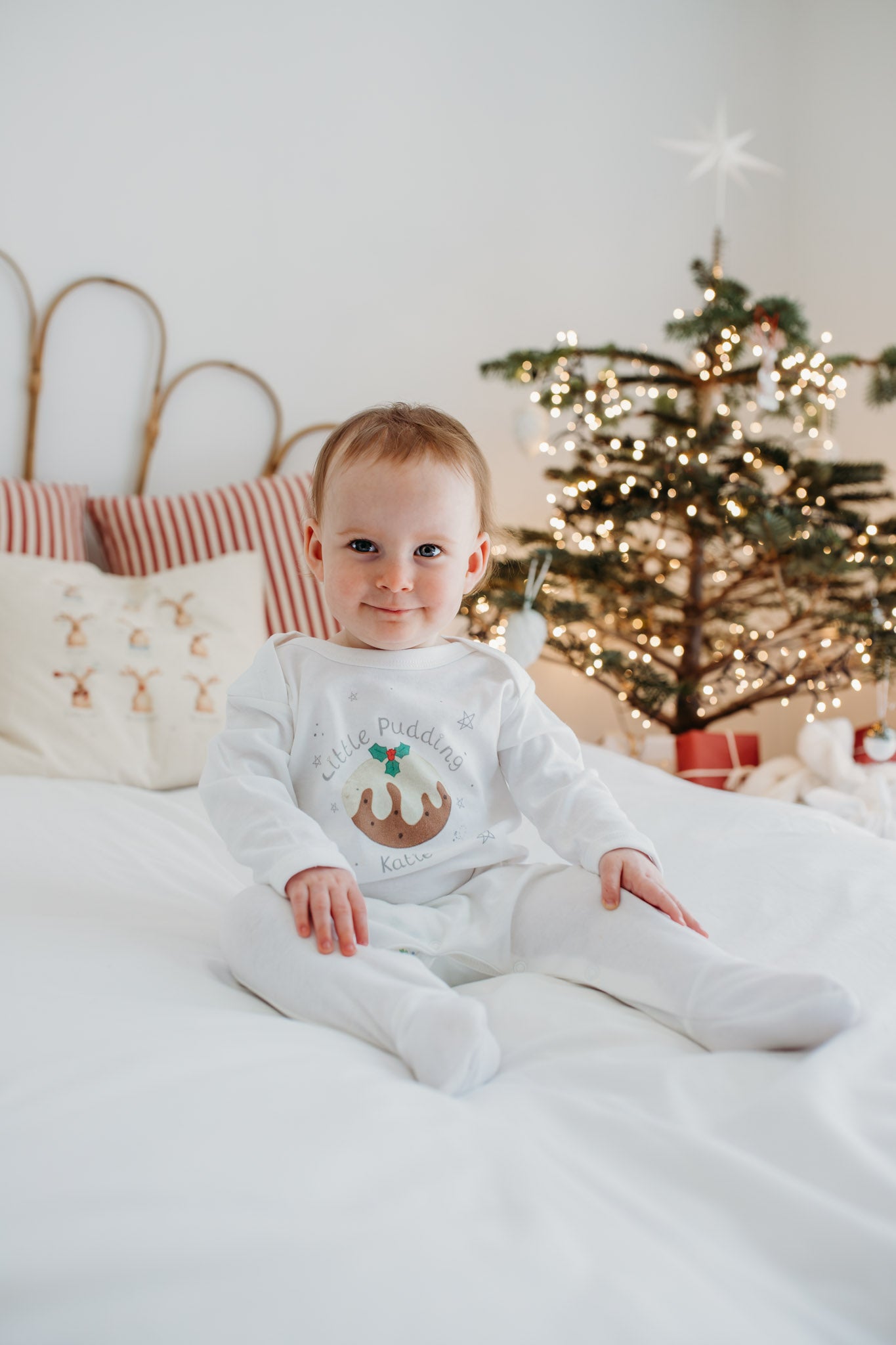 cute baby sat on a bed in front of a christmas tree with sparkling lights wearing a little pudding onesie