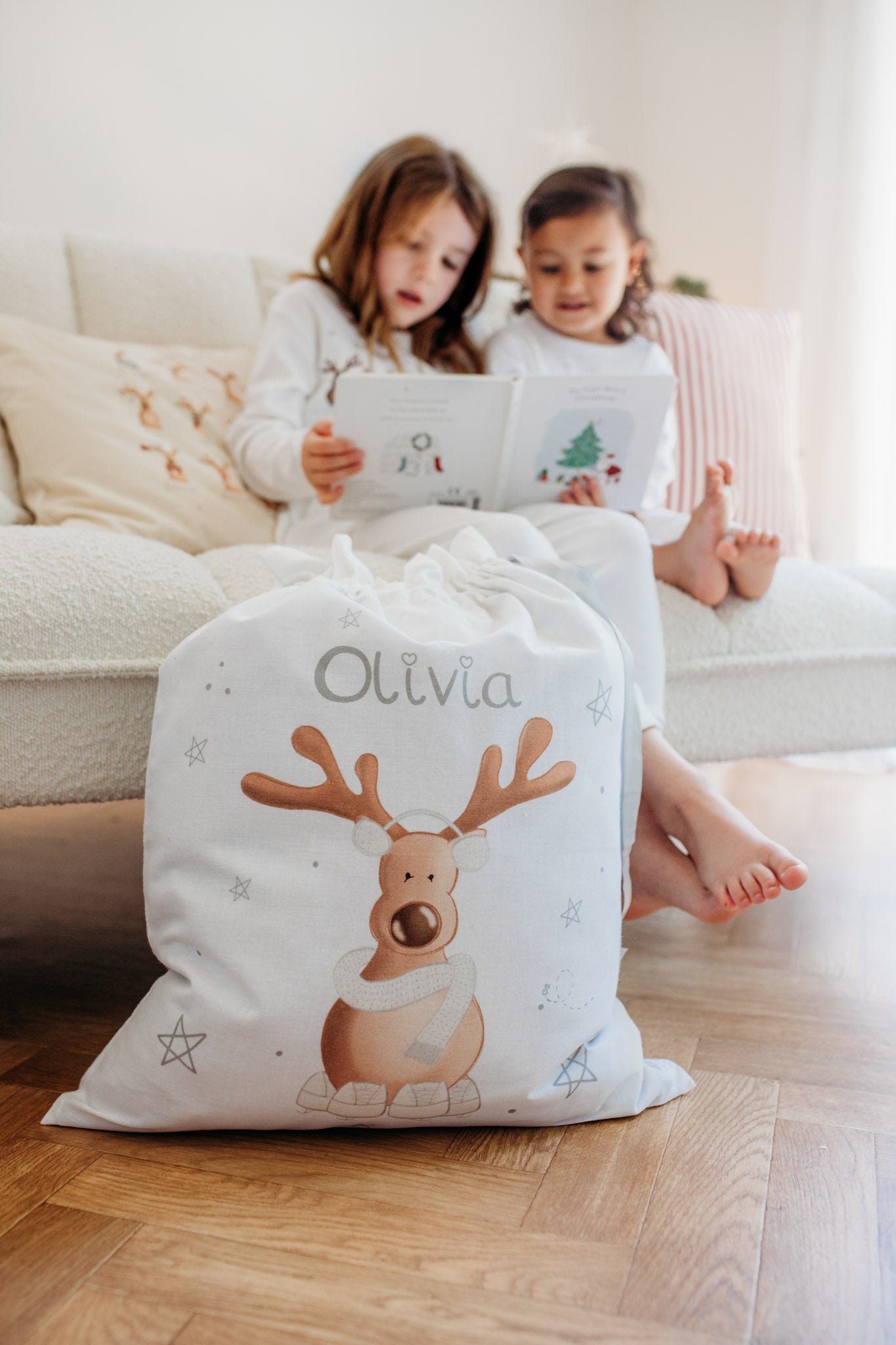 two girls sat on a sofa reading a christmas book with a christmas sack next to them