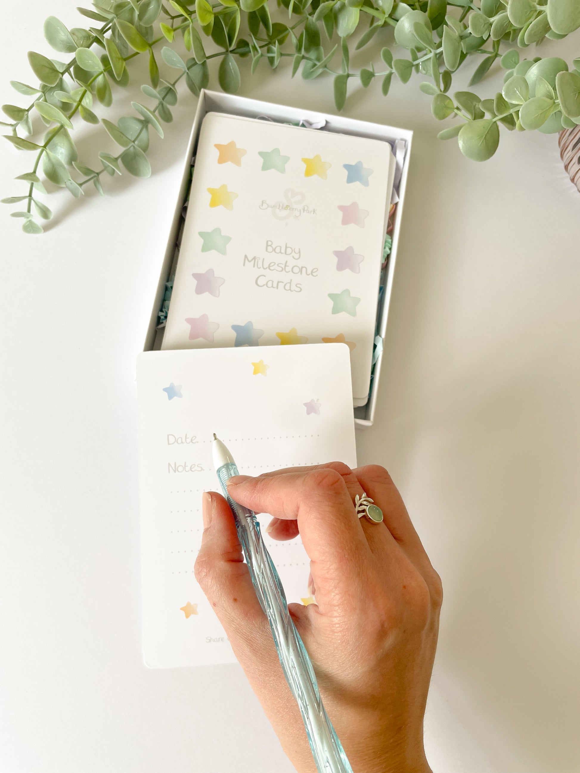 luxury gift set of baby milestone cards in a white gift box and a mummy holding a pen writing keepsake notes on the note card