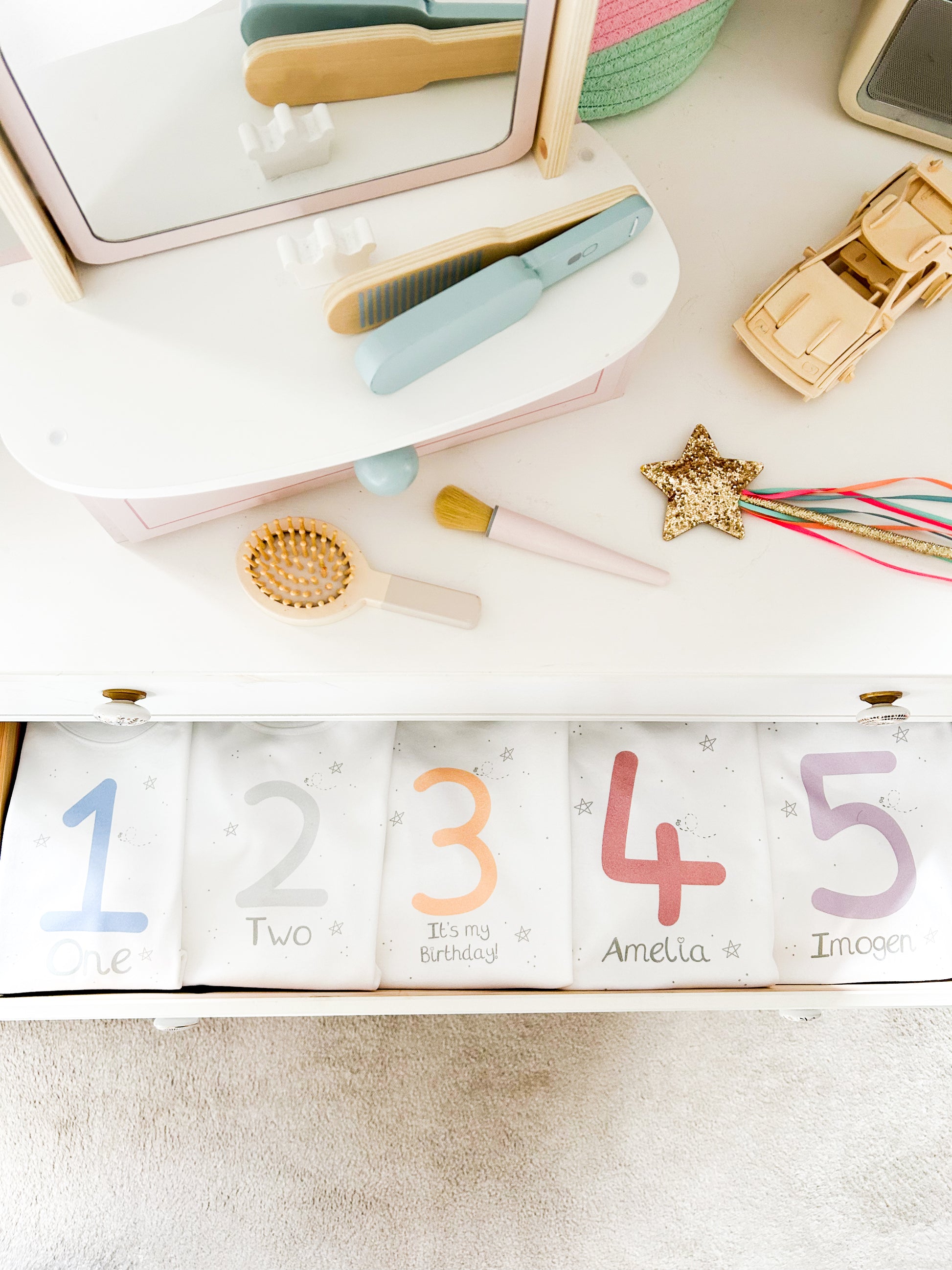 girls bedroom with wooden vanity mirror toy set, magic wand and hair brush with open drawer filled with birthday party clothes personalised with ages for 1st 2nd 3rd 4th and 5th birthday parties in pink, purple, silver, blue and peach and natural colours