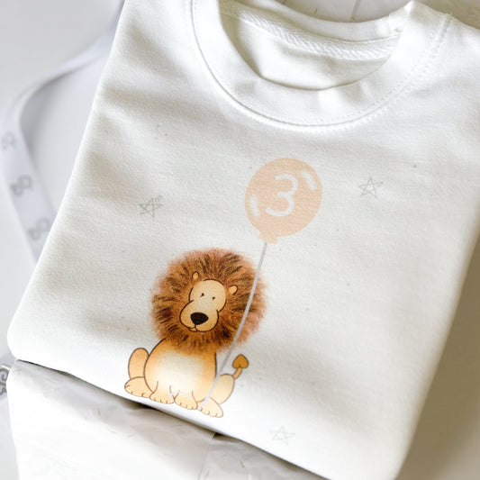 pantone peach fuzz personalised birthday sweatshirt for toddler 3rd birthday party with jungle safari animal lion print holding a party balloon