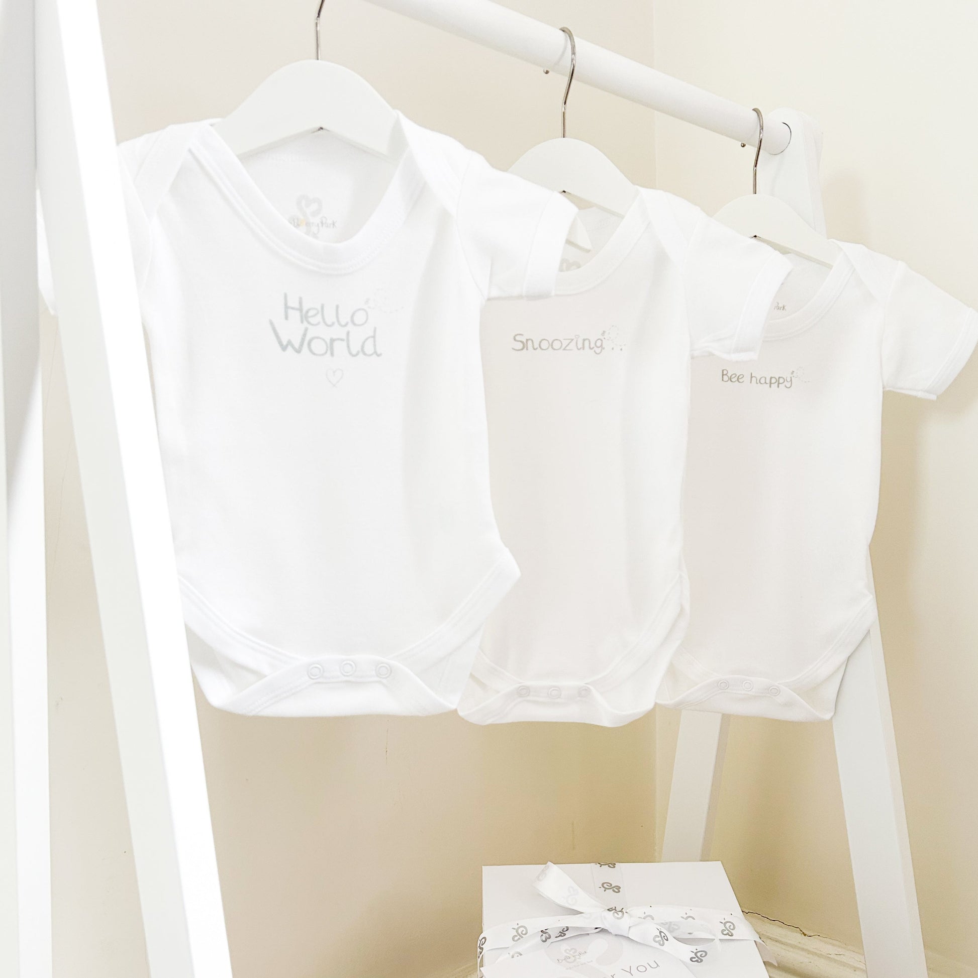 set of 3 white baby bodysuits with silver text prints "hello world" "snoozing" and "Bee happy" on a child's white clothes rail in a children's bedroom