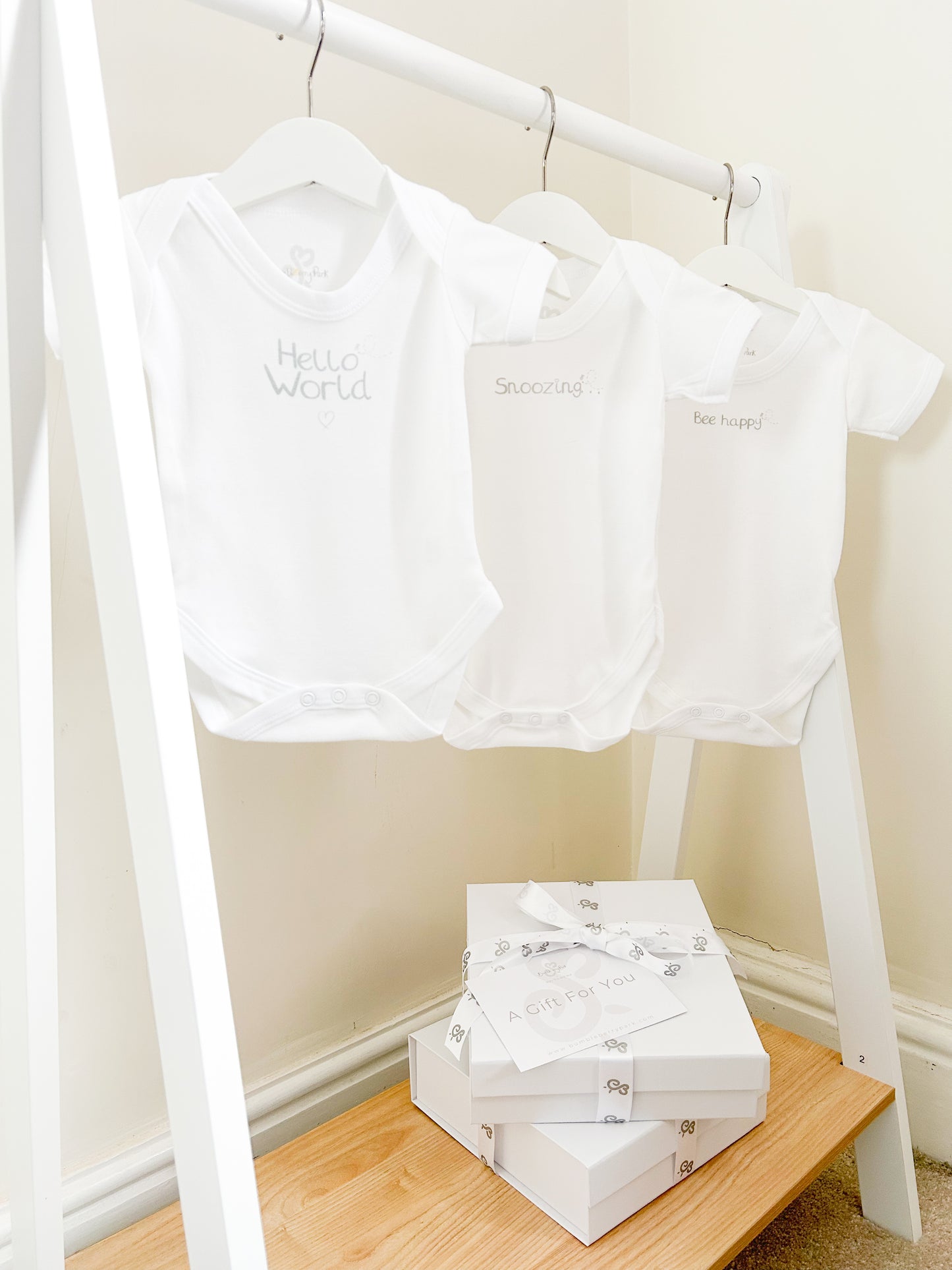 set of 3 mixed size white baby bodysuits with silver text prints "hello world" "snoozing" and "Bee happy" on a child's white clothes rail in a children's bedroom