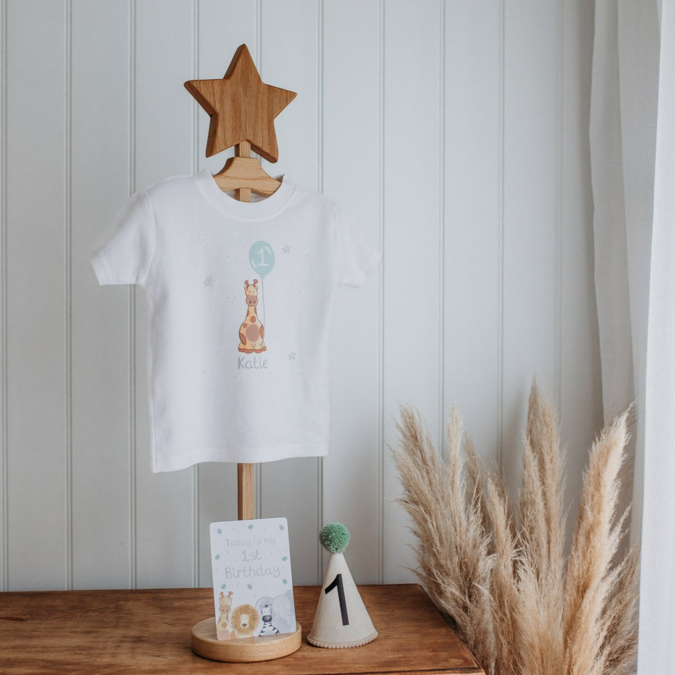 personalised 1st birthday t-shirt with a safari giraffe print holding a party balloon and number 1 on a clothes hanger with a number 1 first birthday party hat and baby milsetone card