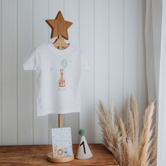 personalised 1st birthday t-shirt with a safari giraffe print holding a party balloon and number 1 on a clothes hanger with a number 1 first birthday party hat and baby milsetone card