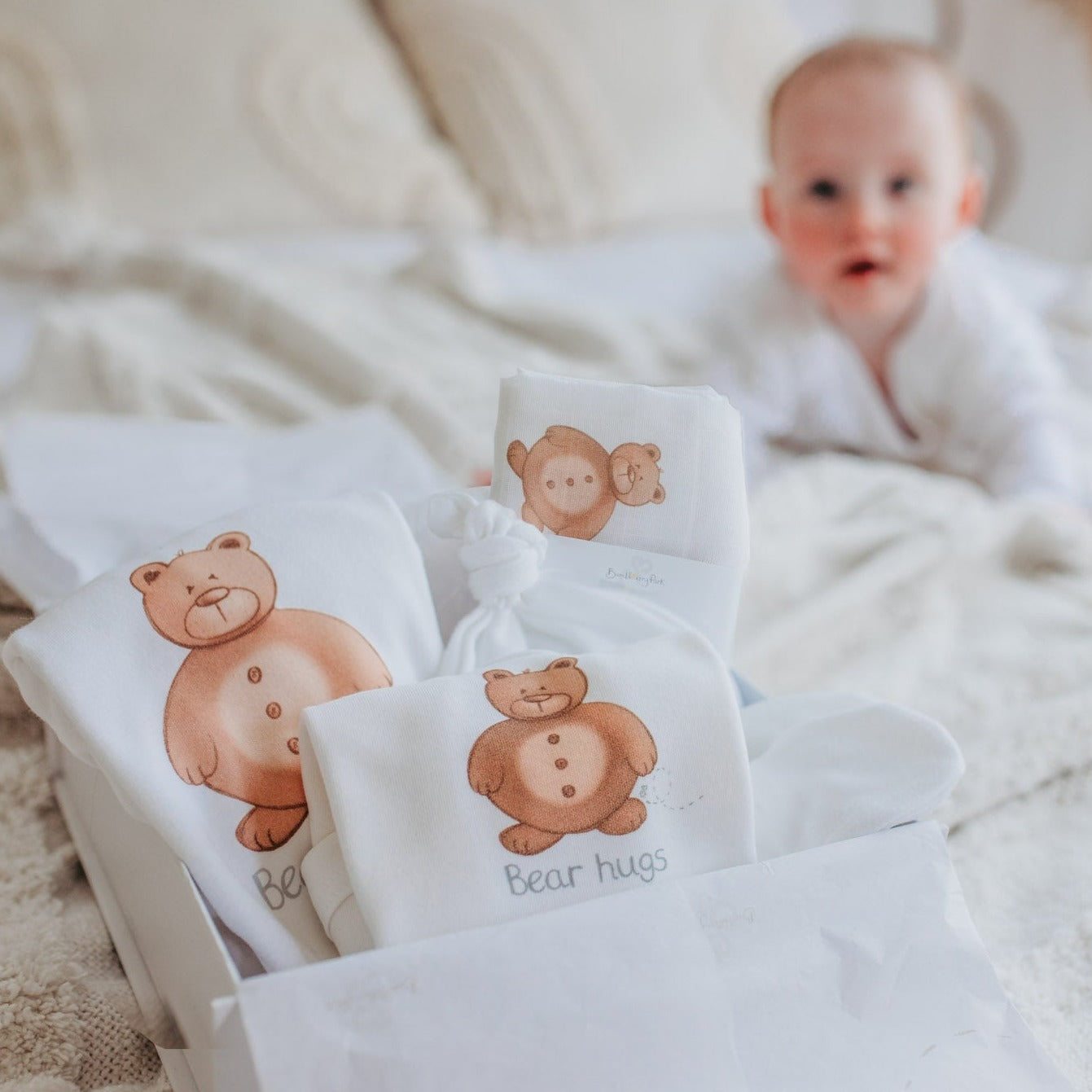 baby laying on a bed next to a new mum and baby gift hamper filled with an organic cotton baby bodysuit, muslin square, sleepsuit, hat and mittens starter set